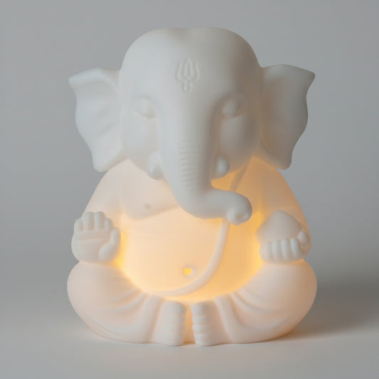 A white Ganesha figurine glowing with warm yellow light from within, placed on a reflective orange-brown surface against a neutral indoor background.
