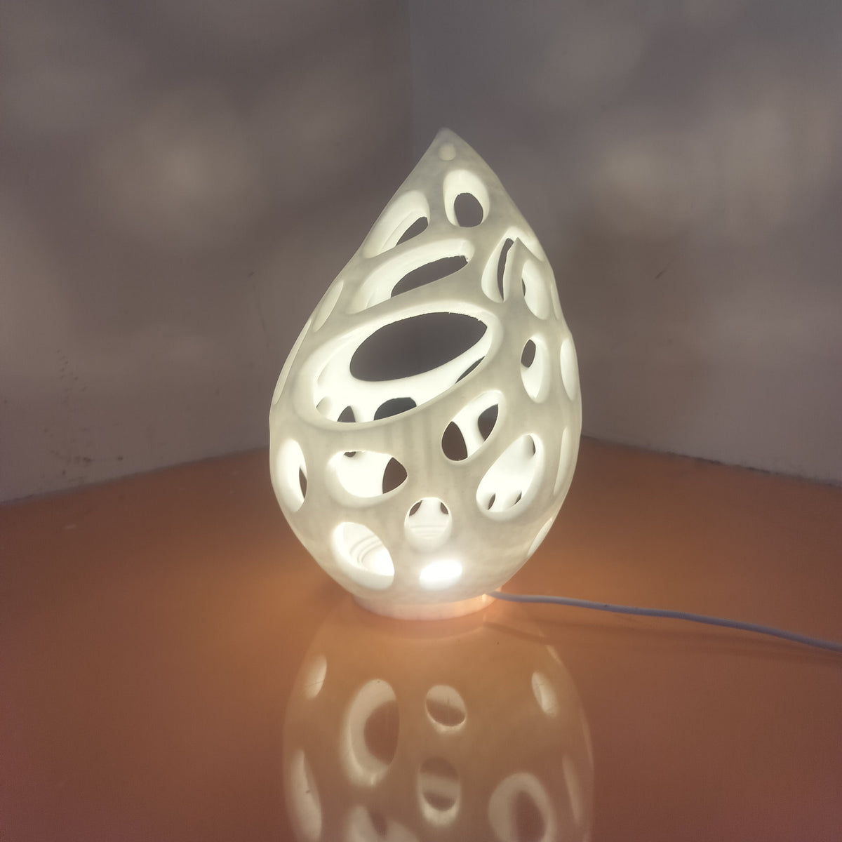 A teardrop-shaped white ceramic lamp with organic cutout patterns, glowing with warm light on a reflective surface against a neutral background.