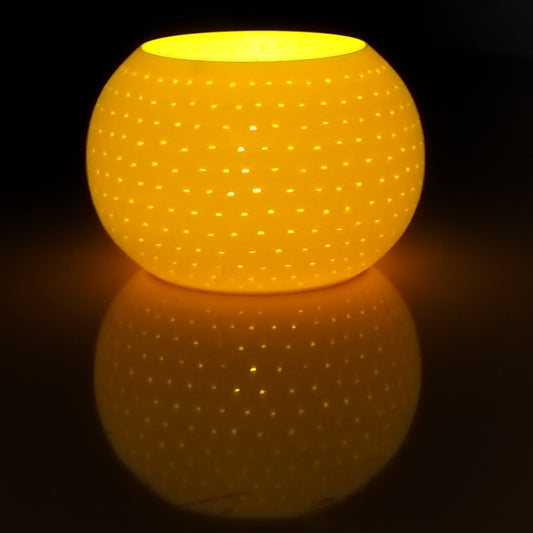 A glowing round yellow-orange lamp or candle holder with a perforated dot pattern, casting a warm reflection on a dark surface against a black background.