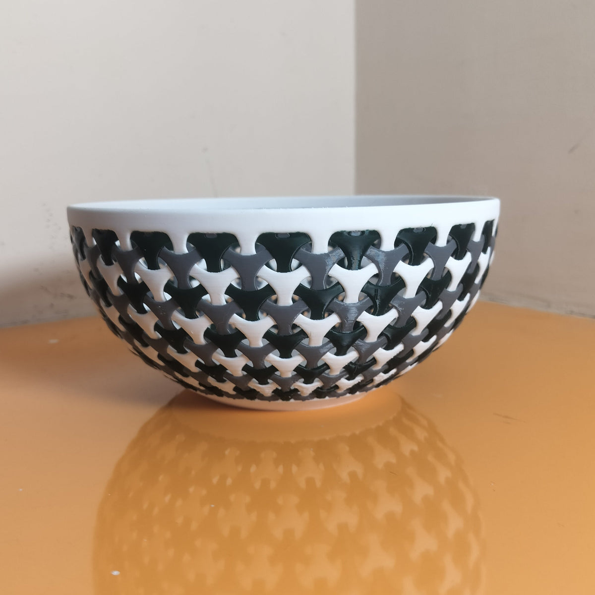 A white ceramic bowl with a black geometric trellis pattern, resting on a reflective orange surface against a neutral beige background.