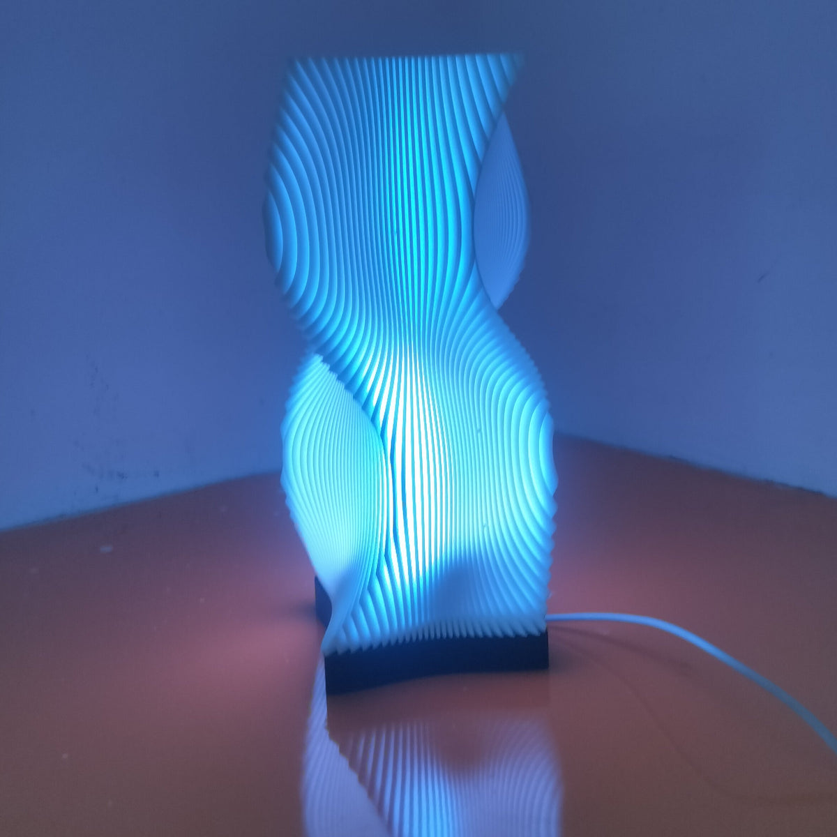 A modern sculptural table lamp with a glowing blue light, featuring a ribbed, wavy vertical design on a dark base, set on a wooden surface in a dimly lit room.