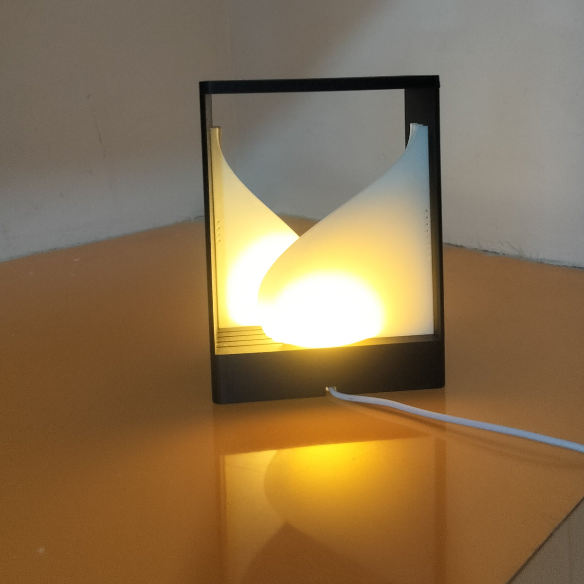 A modern black-framed table lamp with two curved white diffusers glowing warm yellow light, placed on a reflective orange-brown surface against a plain beige wall.