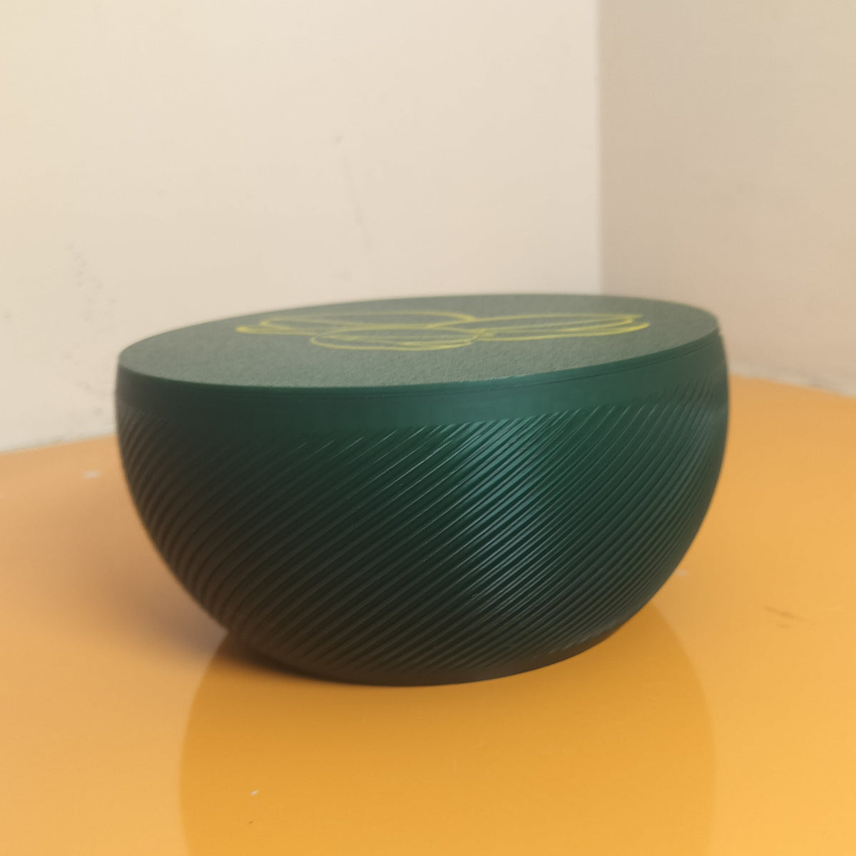 A dark green, ribbed spherical speaker or decorative container with a yellow graphic on top, sitting on a yellow surface against a plain beige wall.