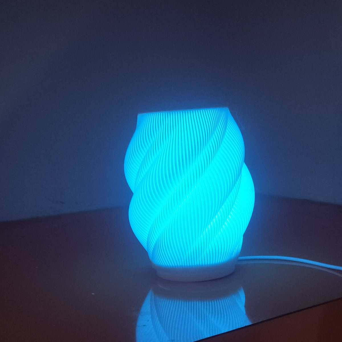 A glowing blue ribbed cylindrical lamp sits on a dark wooden table against a dim wall, with its power cord visible.
