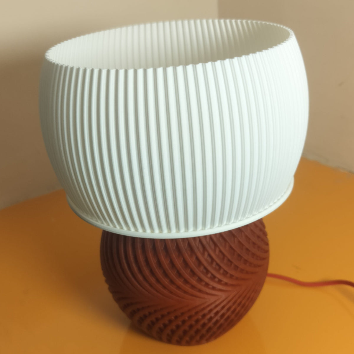 A modern table lamp with a ribbed white cylindrical shade and a textured reddish-brown spherical base, sitting on a yellow-orange table against a beige wall.