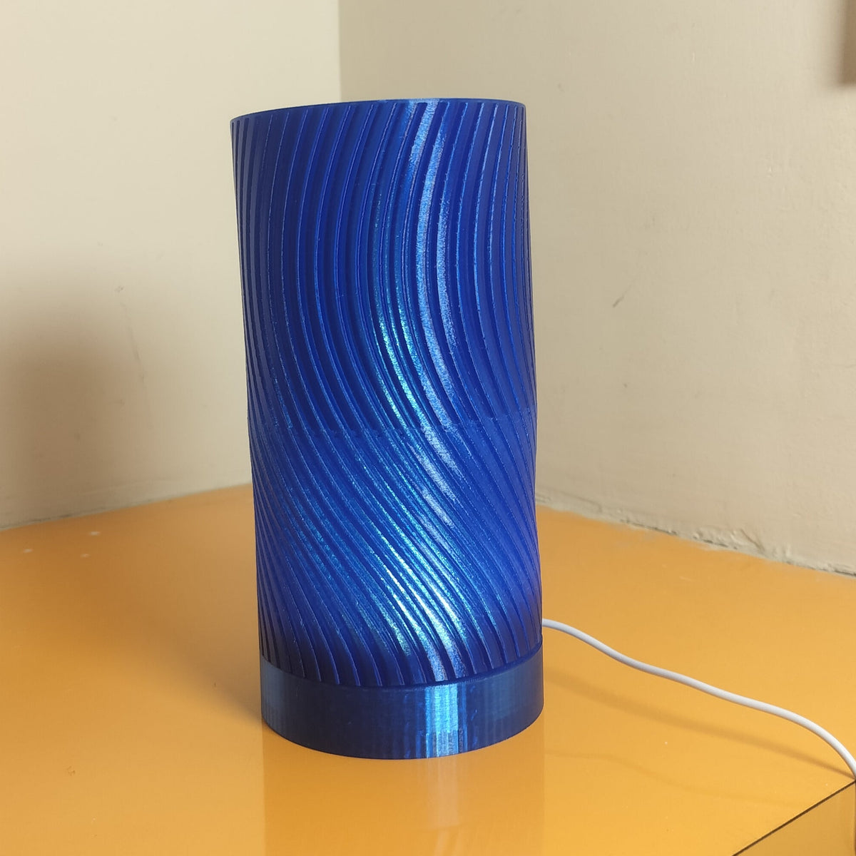 A blue cylindrical lamp or speaker with a glossy, swirled ribbed texture sits on a yellow-orange desk in an indoor room setting.