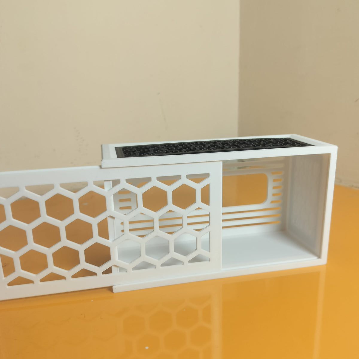 A white 3D-printed modular screen panel with a honeycomb pattern and ventilation slots, shown on a reflective orange surface against a beige wall.