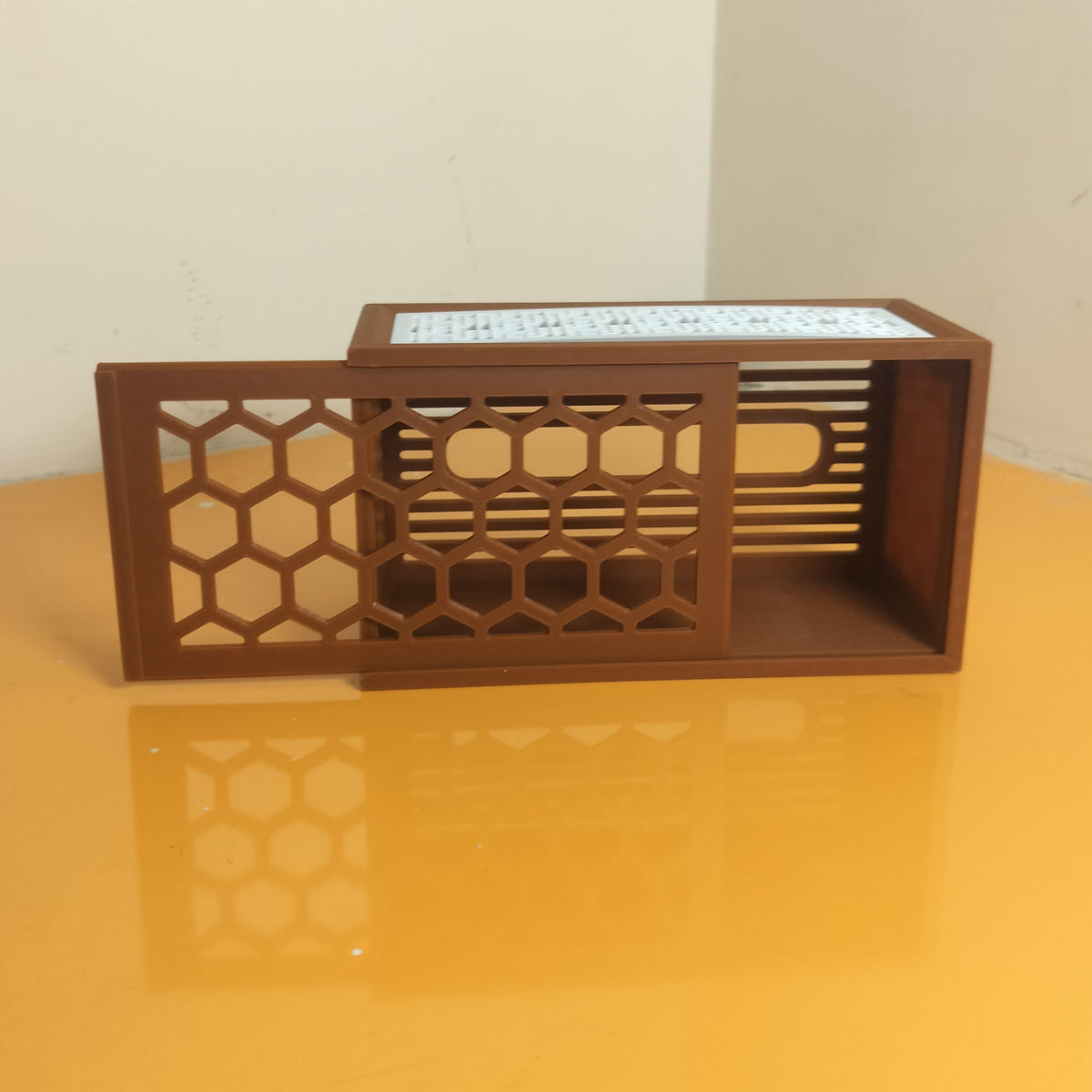 A small wooden box with a sliding honeycomb-patterned door, placed on a yellow reflective surface against a plain off-white wall.