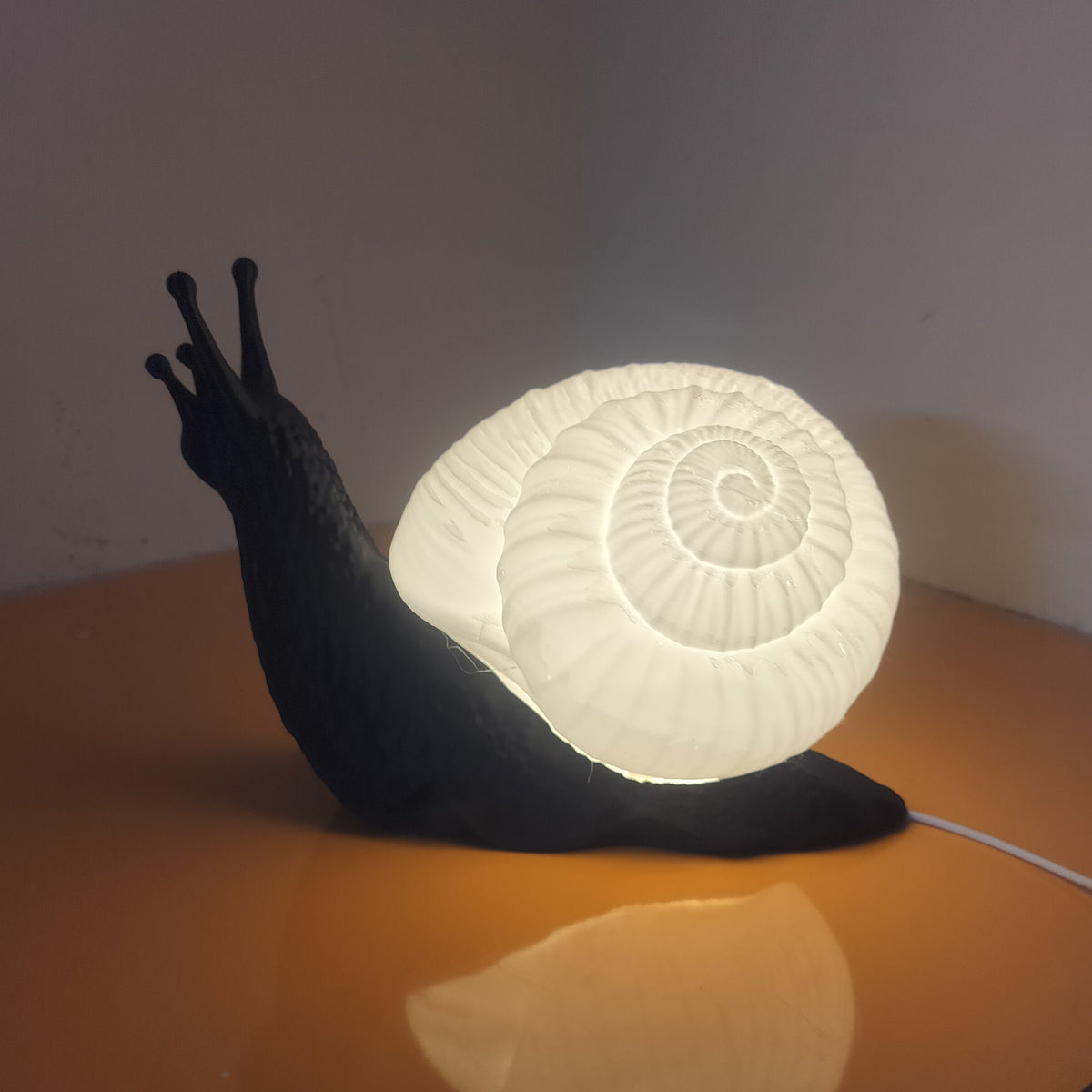 A snail-shaped table lamp with a glowing white spiral shell and a dark body, placed on a reflective orange surface in a dim room corner.