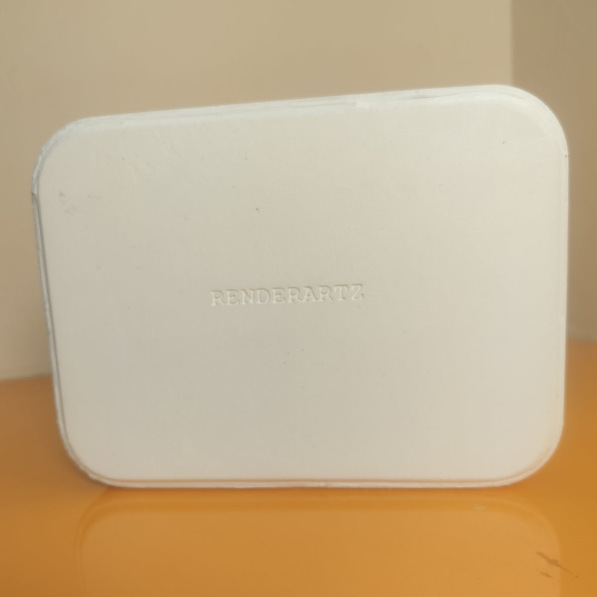 A white rectangular compact case with rounded corners and the embossed brand name 'RENDE PARTS' in the center, resting on a reflective orange surface against a beige background.