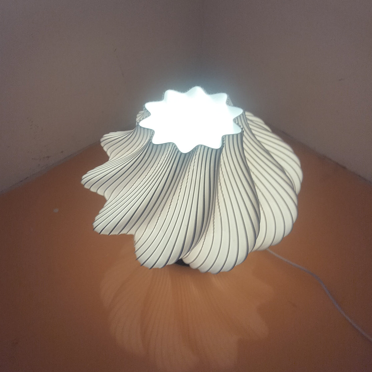 A modern sculptural table lamp with a ribbed, flower-like shade that glows with a cool white light from within, placed on a brown surface in a room corner.