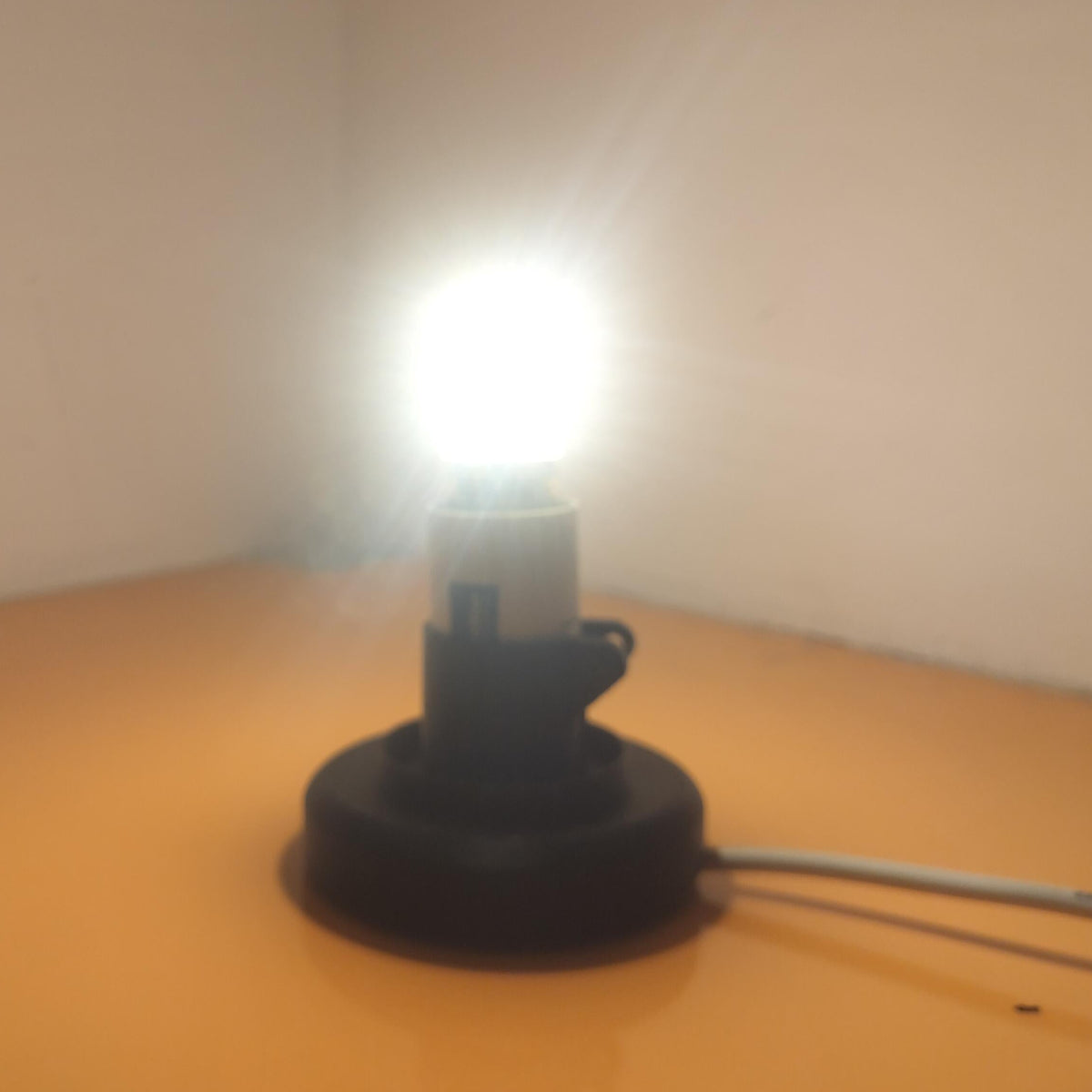 A small black floor lamp with a bright glowing bulb, set on a glossy orange surface against a plain beige wall corner.