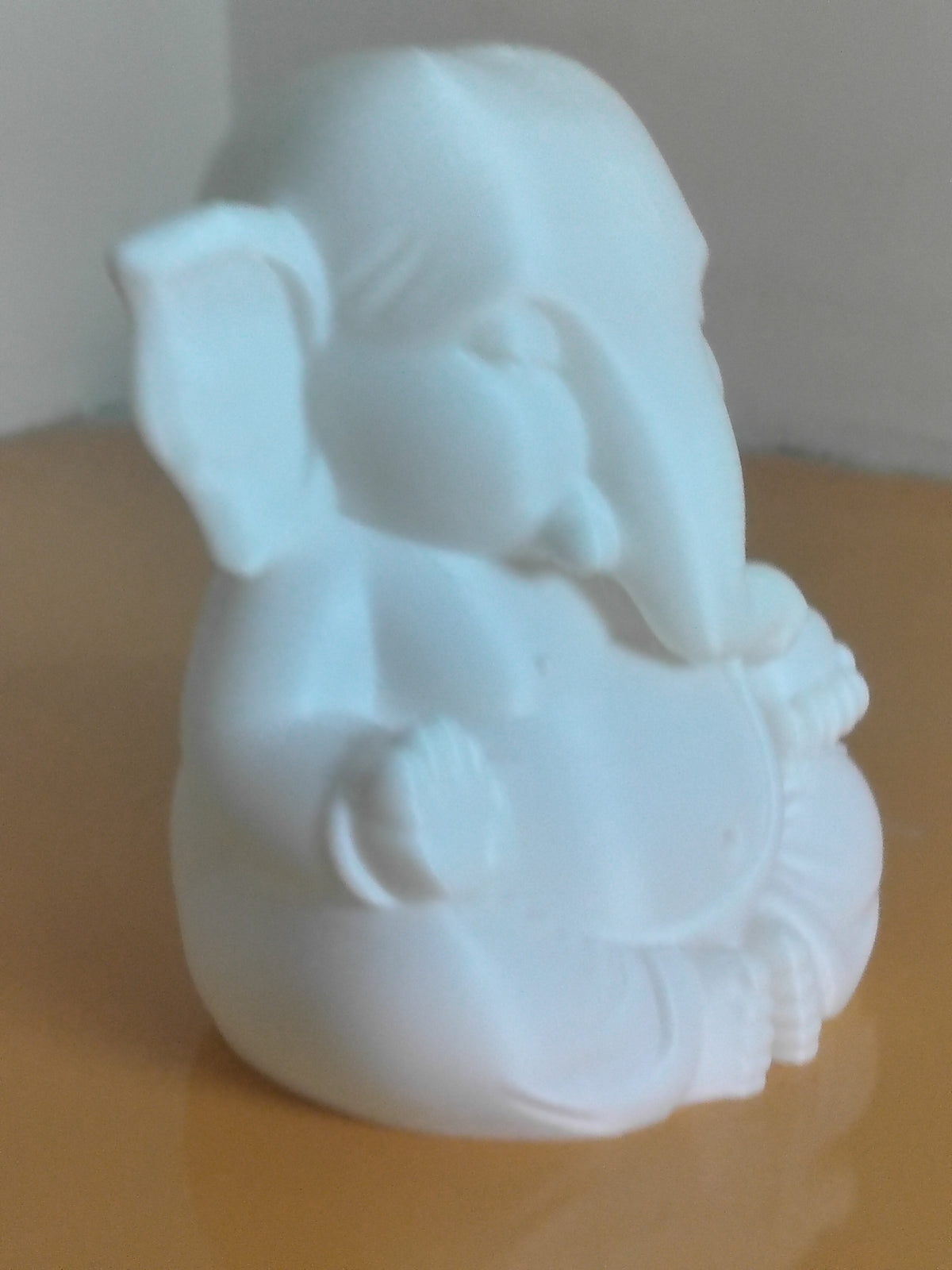 A small white Ganesha, likely made of ceramic or stone, shown sitting on a warm beige reflective surface against a neutral light gray background.