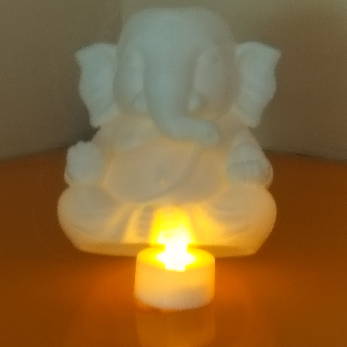A white Ganesha figurine glowing with warm light, positioned behind a small white tea light candle on a reflective orange surface.