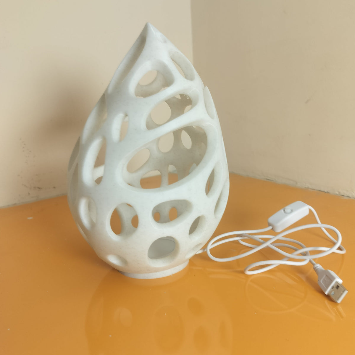 A white 3D-printed teardrop-shaped LED light with a perforated, lattice-like design, shown next to its white USB charging cable on a reflective orange surface.