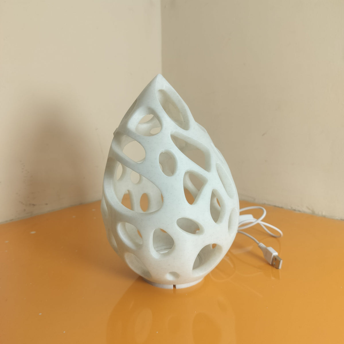 A white 3D-printed teardrop-shaped lamp with an organic, open lattice pattern, sitting on a reflective orange surface against a beige wall corner.