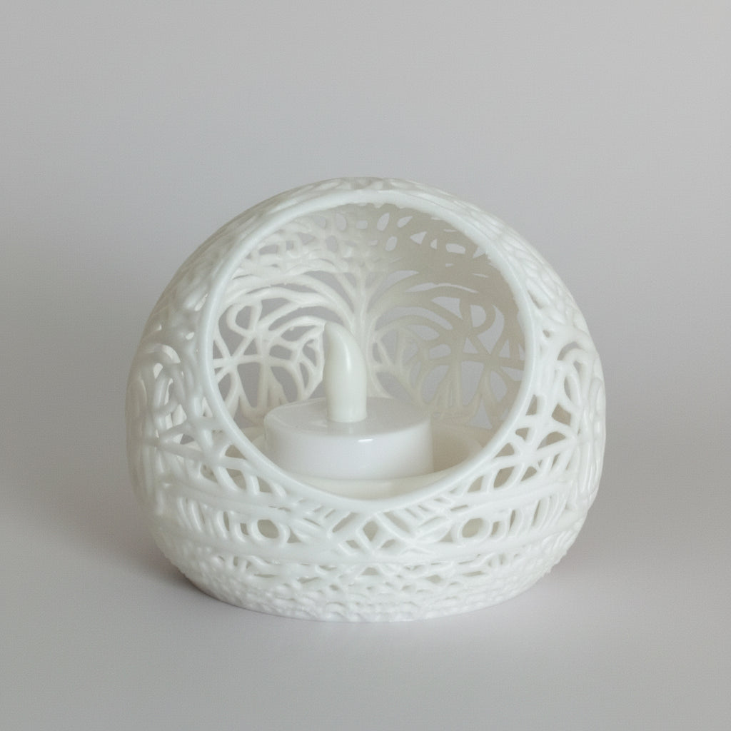 A white spherical laser-cut candle holder with an intricate tree design, containing a small white LED tea light candle, set on a reflective orange surface against a beige background.