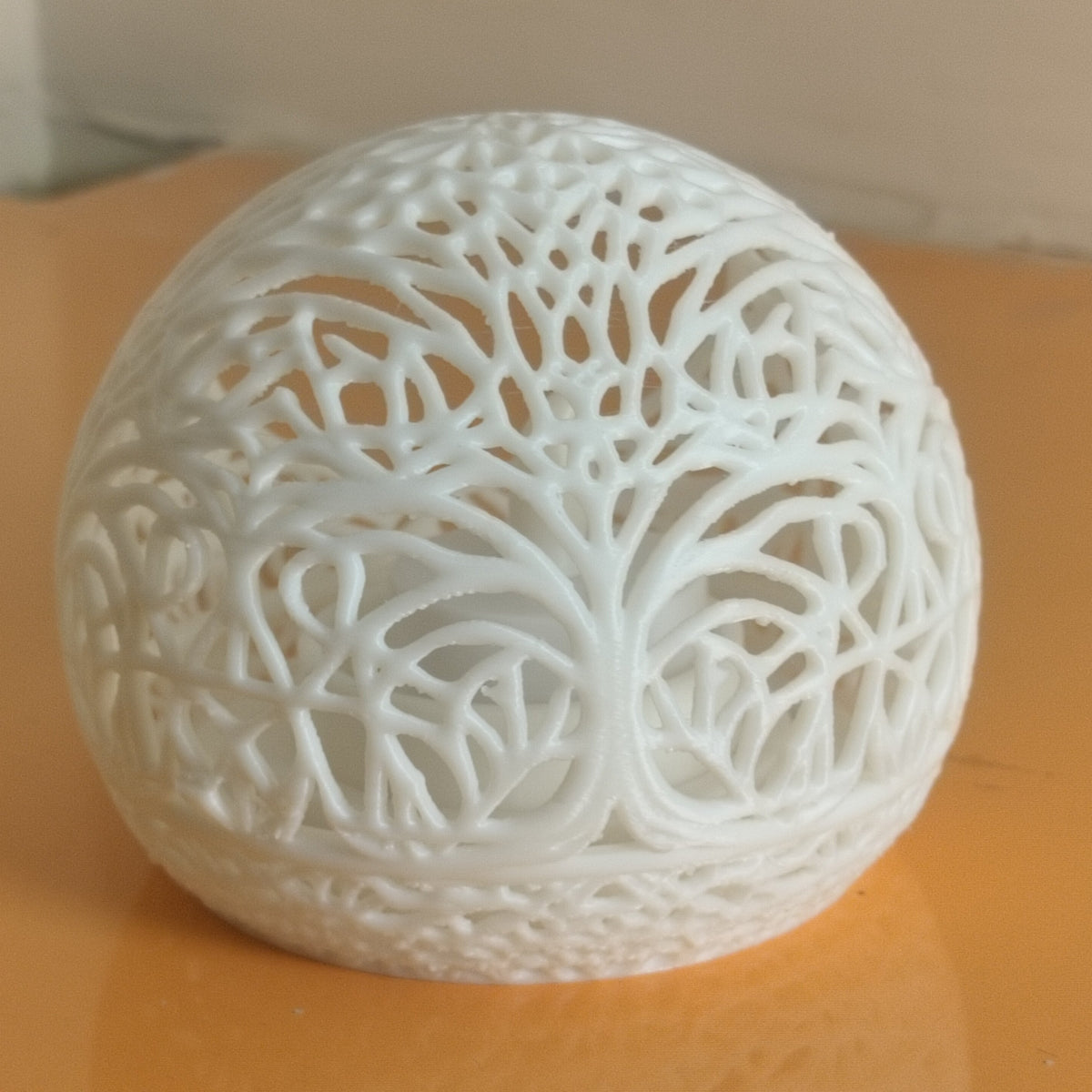 A white 3D-printed decorative sphere with an intricate tree-of-life cutout pattern, resting on a reflective orange surface.