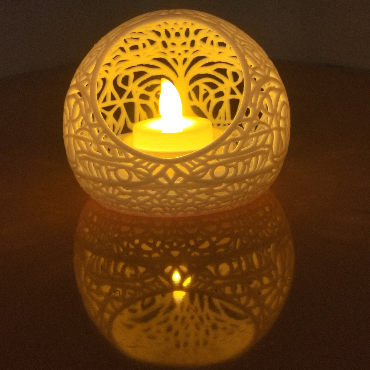A glowing LED tea light candle holder with an intricate laser-cut tree of life pattern, casting a warm yellow light and reflection on a dark surface.