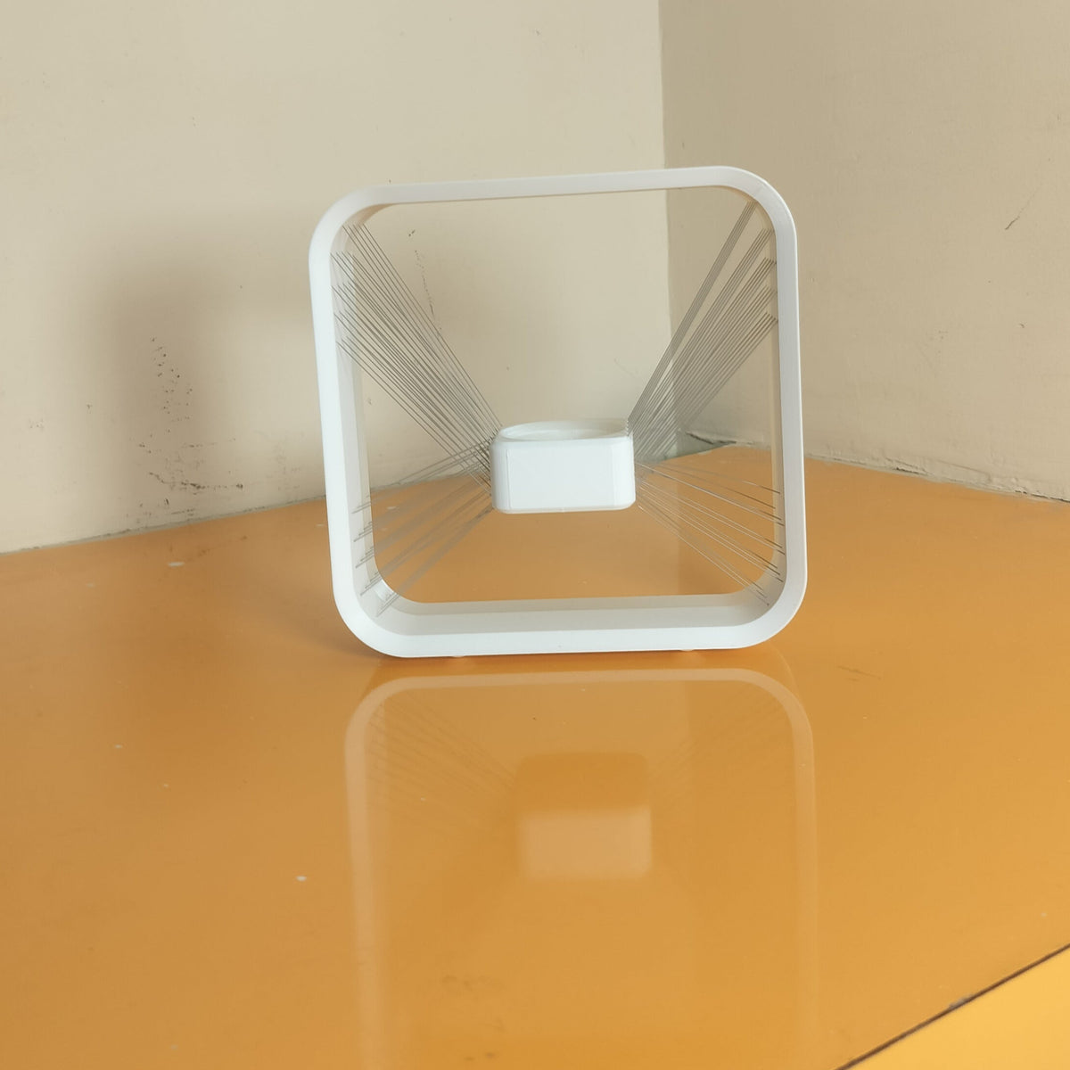 A square white plastic container with a clear glass insert, standing on a yellow-orange surface against a beige wall.