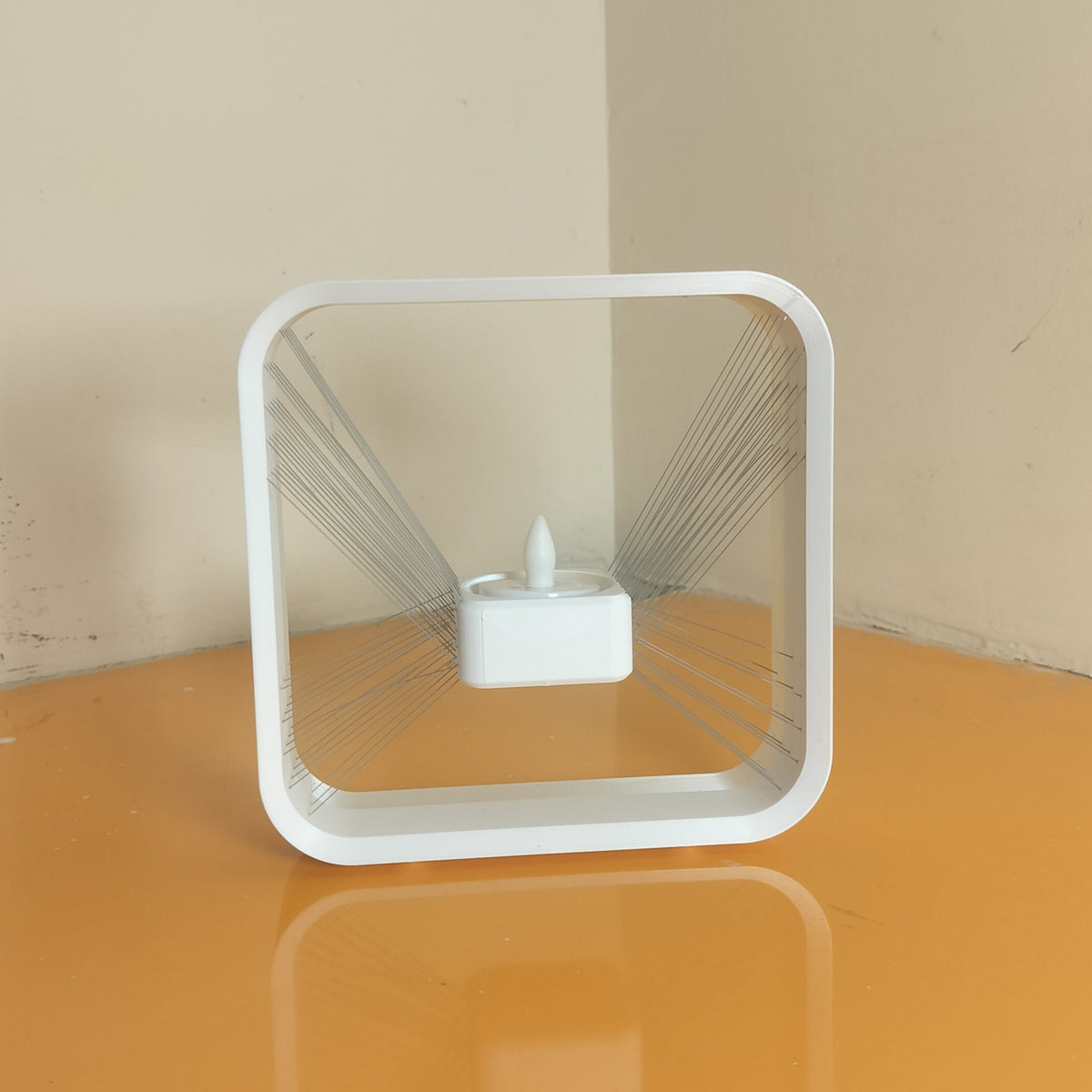A white square LED light strip with a central bulb, standing on a reflective orange surface against a beige wall corner.