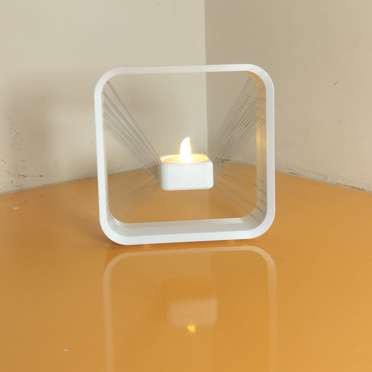 A modern square white LED tea light candle holder with a clear acrylic front panel, displaying a small lit candle, set on a glossy orange table against a beige wall.