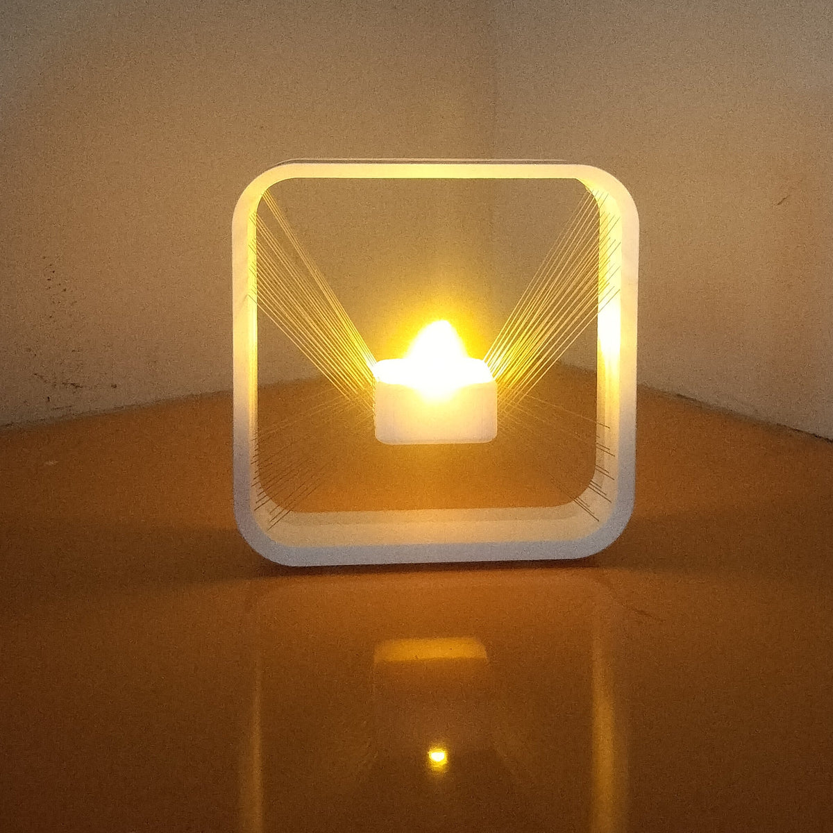 A glowing square LED light box with a small candle inside, sitting on a reflective brown surface against a dimly lit gray wall.