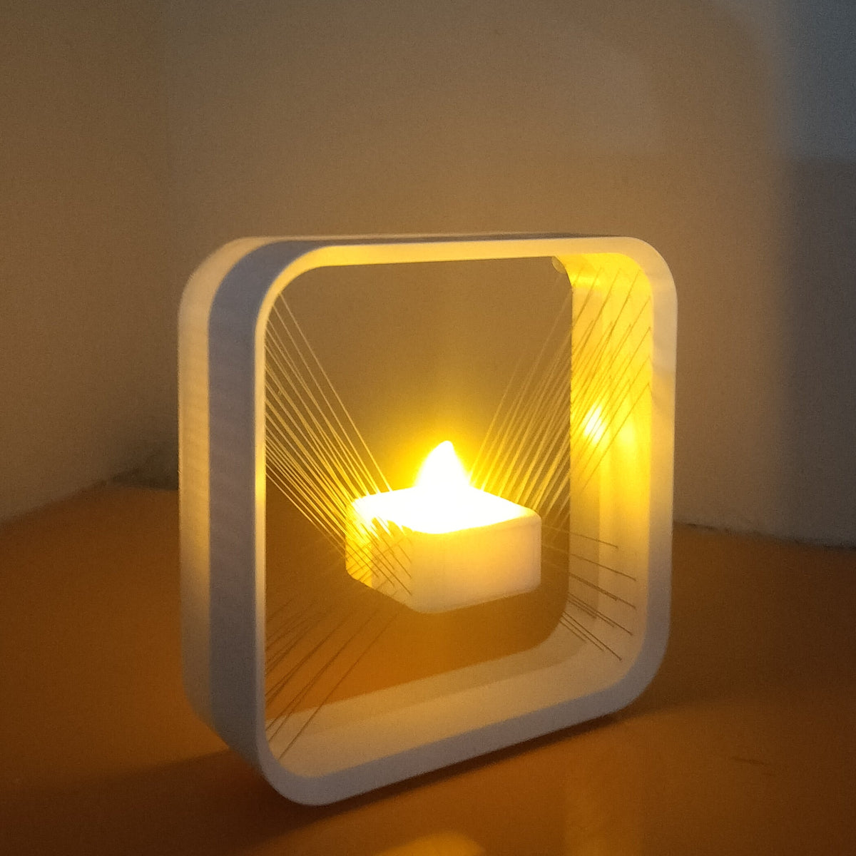 A modern square white LED candle holder lamp with a glowing yellow flame effect, sitting on a reflective brown surface against a plain wall.