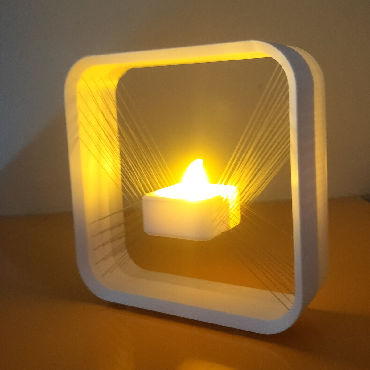 A modern square LED light box with rounded corners, featuring a glowing yellow flame effect and a small white candle-like element inside, set against a dark background.