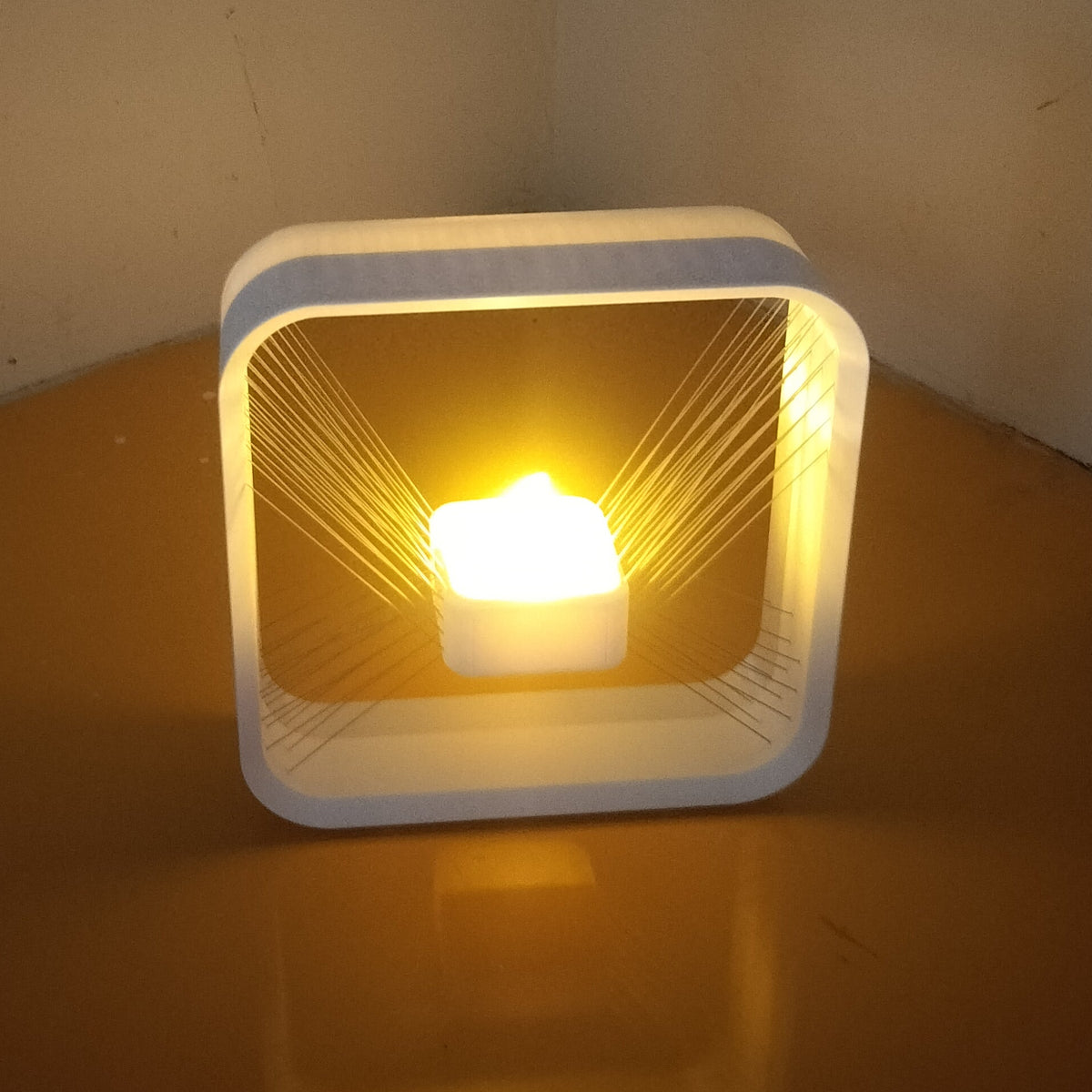 A square white LED candle-style night light with a glowing warm yellow light inside, placed on a wooden surface against a plain wall.