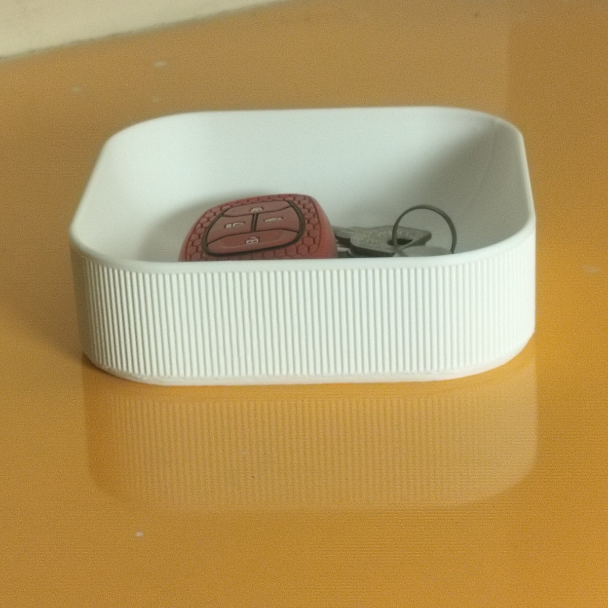 A square white  3D printed PLA catchall tray with a ribbed side, holding a red and black car key fob, placed on a yellow-orange reflective surface.