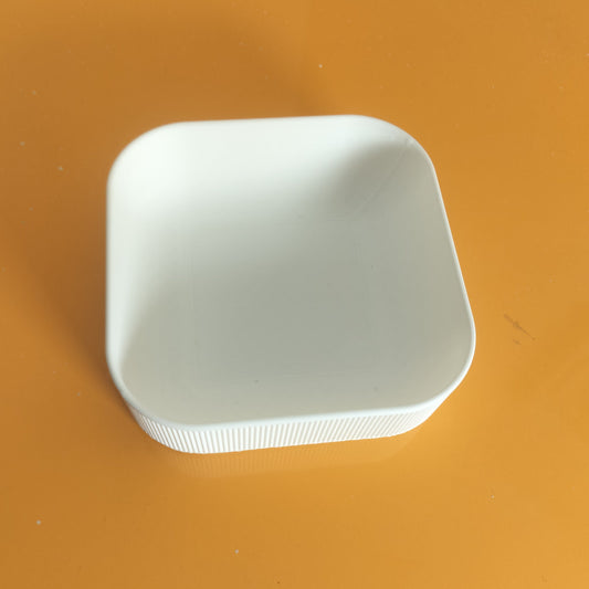 A small white square ceramic dish with rounded corners and a ribbed outer edge, placed on a glossy orange surface.