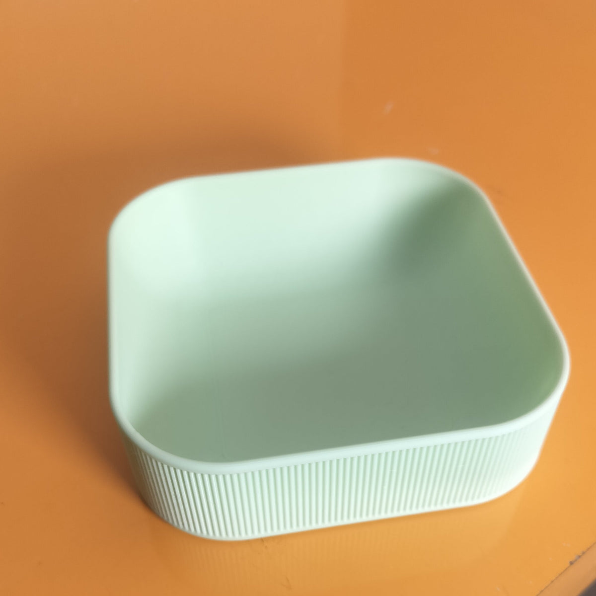 A square, mint green plastic container with a ribbed exterior and rounded corners, shown on a glossy orange table.