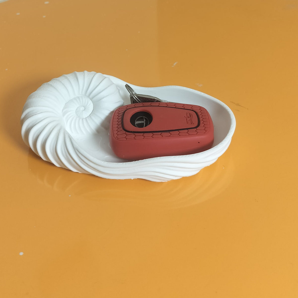 A red car key fob resting on a white, ridged circular coaster placed on a glossy orange surface in the corner of a room with beige walls.