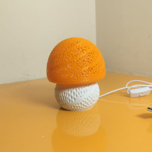 A mushroom-shaped lamp with a textured orange cap and a white textured base, sitting on a reflective yellow-orange table with its white power cord visible.