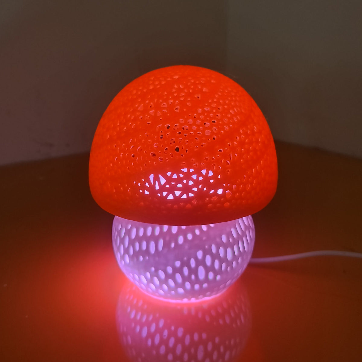 A mushroom-shaped LED table lamp with a glowing orange-red cap and a purple-white perforated base, sitting on a dark surface against a dim wall.