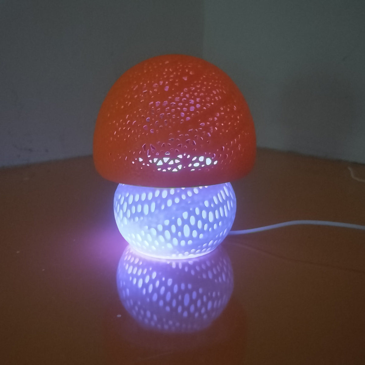 A mushroom-shaped decorative lamp with a textured orange cap and a glowing white base, casting purple light onto a dark surface.