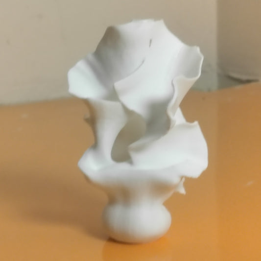 A white ceramic sculptural vase with a flared, organic ruffled rim and a rounded base, standing on a reflective orange surface against a beige wall.