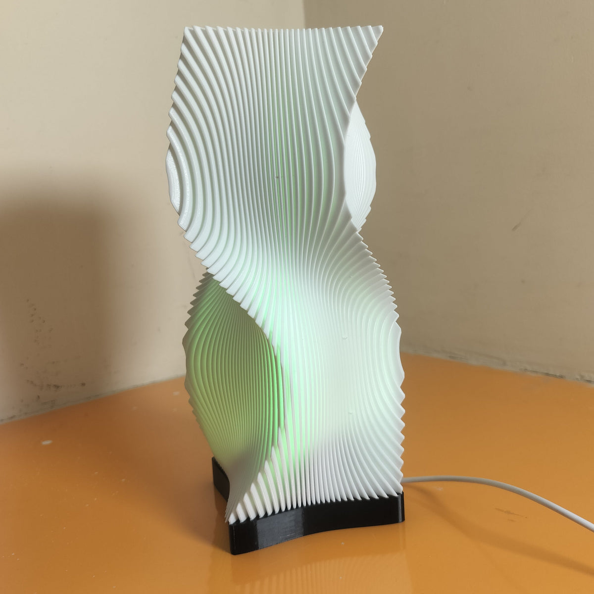 A modern sculptural table lamp with a white, wavy, accordion-like layered design on a black base, emitting a soft green glow, set on an orange surface against a beige wall.