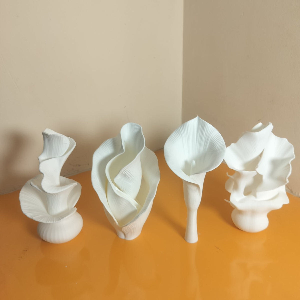 Four white calla lily flower sculptures of varying heights and designs, arranged on a glossy orange surface against a beige wall.