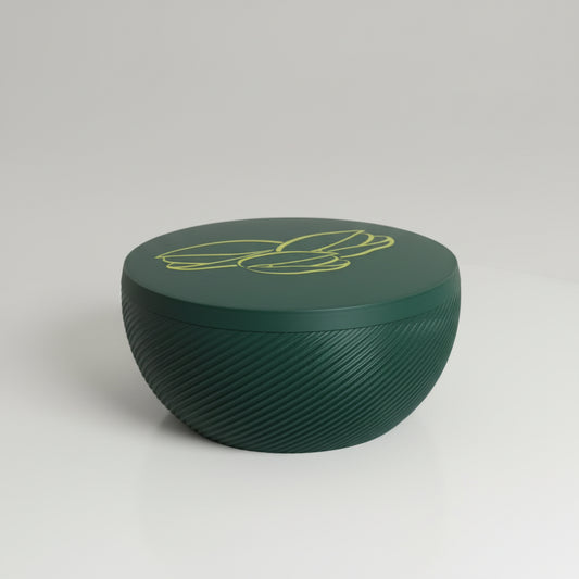 A dark green circular textured container with a yellow line-art illustration of leaves on top, sitting on a yellow-orange surface against a beige wall corner.