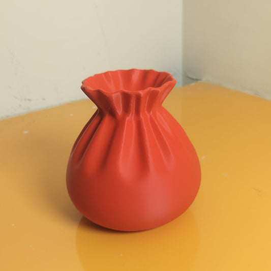 A matte red ceramic vase with a gathered, drawstring-like top rim, sitting on a yellow tabletop against a plain off-white wall corner.