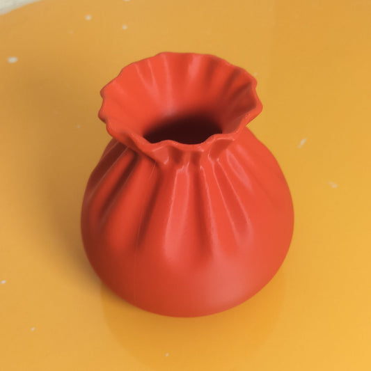A small red fabric pouch vase with a ruffled, gathered opening, placed on a yellow surface in a corner.