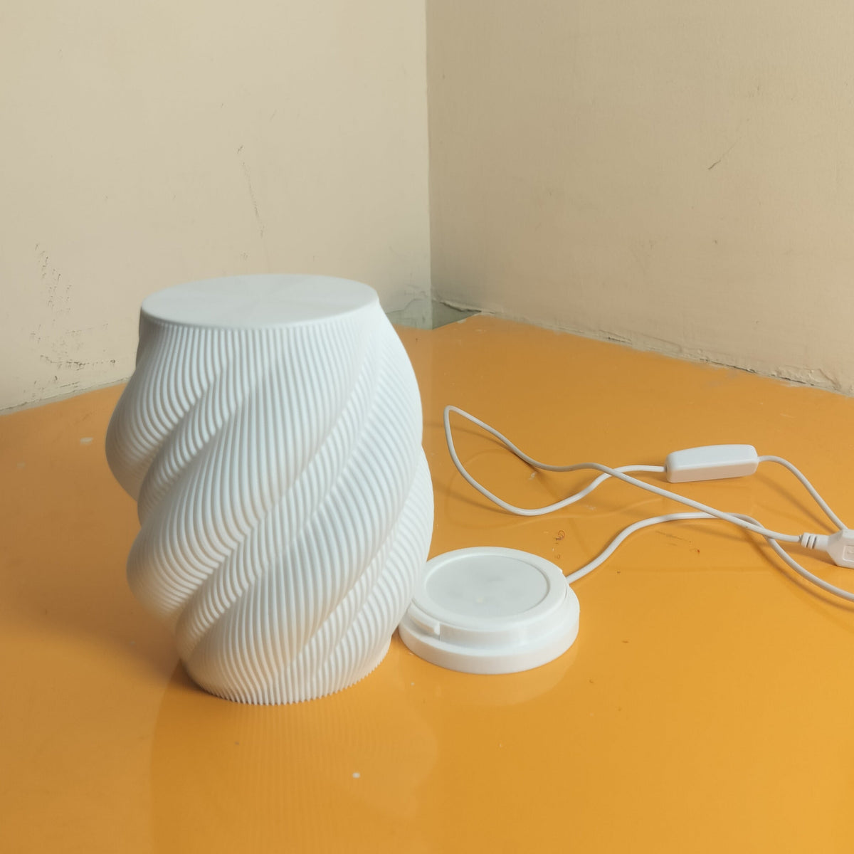 A white ribbed cylindrical lamp or light fixture sits on a yellow-orange table corner, with its power cord and small round accessory nearby against a beige wall.