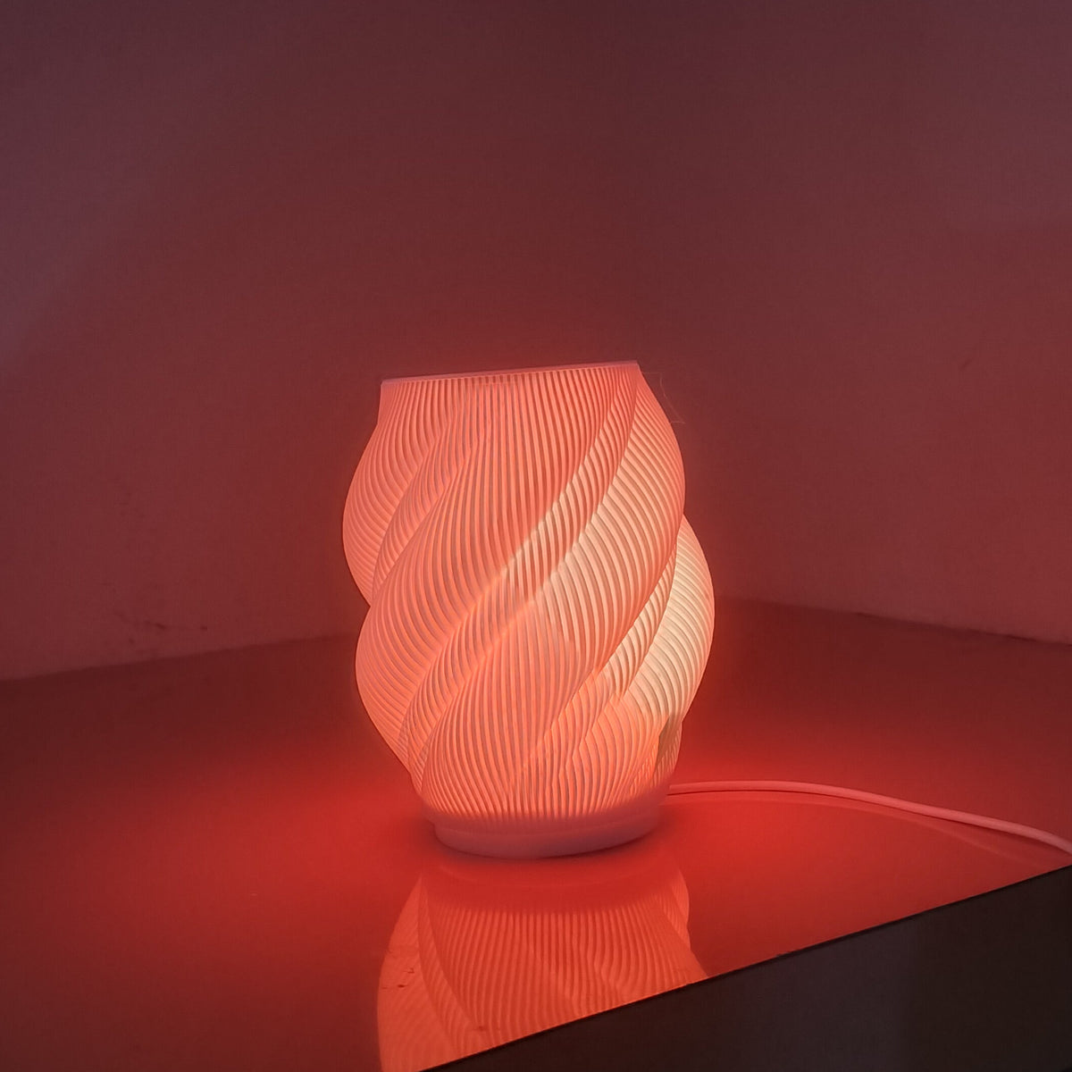 A glowing spiral-patterned lamp emitting a warm red-orange light, placed on a reflective tabletop in a dim room.