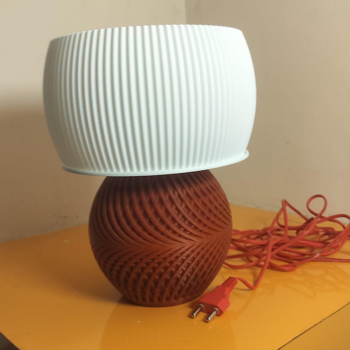 A table lamp with a white ribbed cylindrical shade and a brown textured spherical base, shown on a yellow table with its red power cord in a home setting.