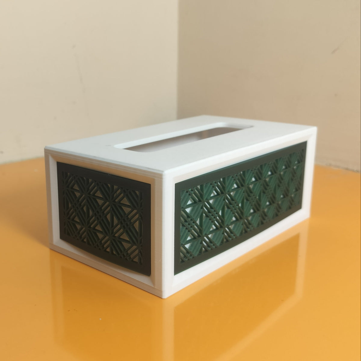 A white rectangular tissue box cover with dark green geometric patterned front panels, placed on a glossy orange surface against a beige wall corner.