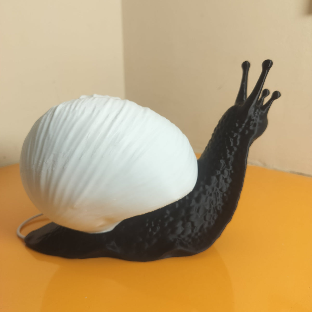 A snail-shaped lamp or figurine with a large white ribbed shell and a black body and antennae, placed on a glossy orange surface against a beige wall.