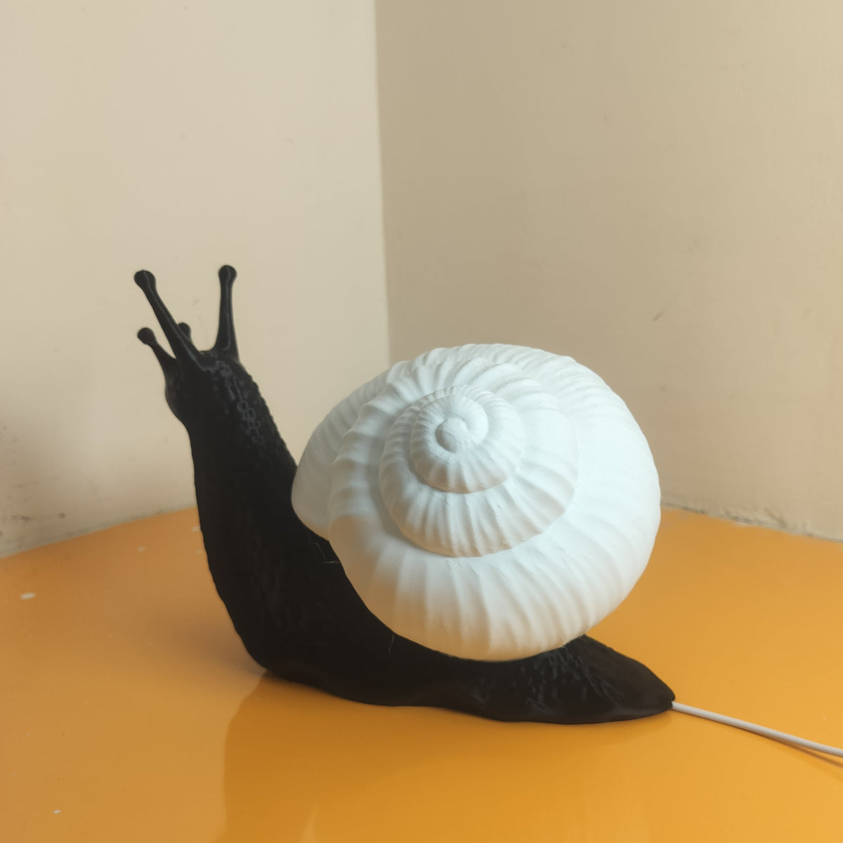 A novelty snail-shaped lamp or figurine with a black body and a large white spiral shell, placed on a glossy orange tabletop against a beige wall.