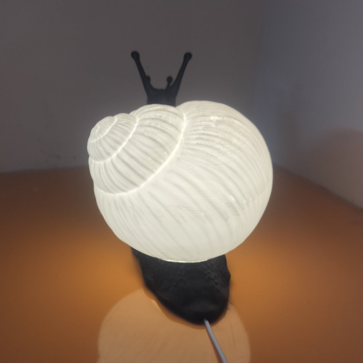 A snail-shaped table lamp with a glowing white shell and a dark body, placed on a brown surface against a gray background.